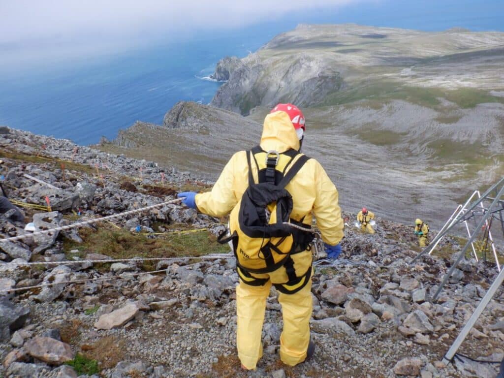 Person in a hazmat suit holding a rope while hiking down a mountain